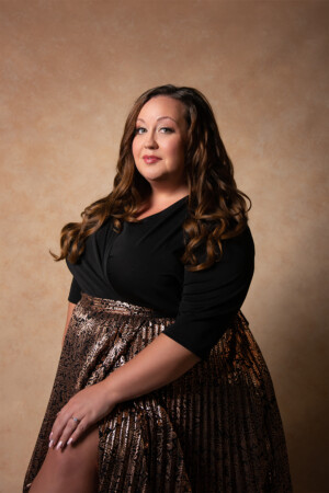 Fine art portrait of curvy woman in black and copper dress seated in front of gold background